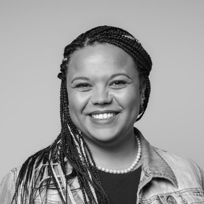 An image of a woman with long dark hair,wearing a pearl necklace, dark t-shirt and light shirt, smiling.