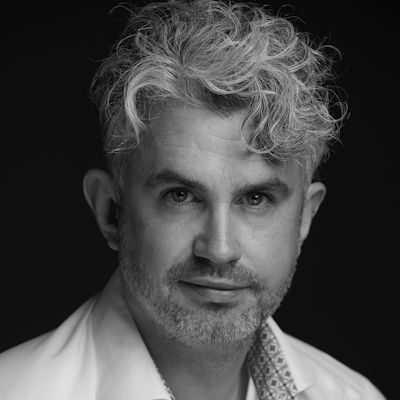A black and white close-up of a man with medium-length light hair and beard, wearing a white shirt.