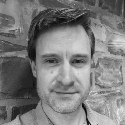 A black and white close-up of a man with short dark hair wearing a light shirt, leaning against a stone wall.