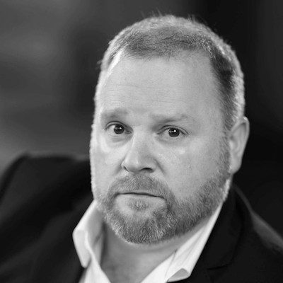 A black and white close-up of a man with short light-coloured hair and a goatee, wearing a white shirt and black jacket.