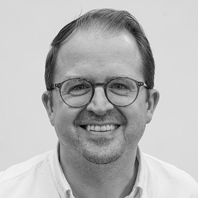 A black and white close-up of a man with dark receding hair, wearing glasses and a white shirt smiling.