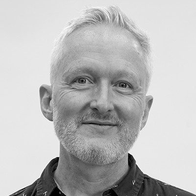 A black and white close-up of a man with short light hair wearing a dark shirt, smiling.