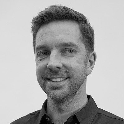 A black and white close-up of a man with short dark hair and stubble wearing a dark shirt.