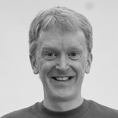 A black and white close-up of a man with short blonde hair wearing a dark t-shirt, smiling.