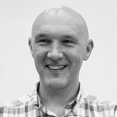 A black and white close-up of a bald man smiling, wearing a chequered shirt.