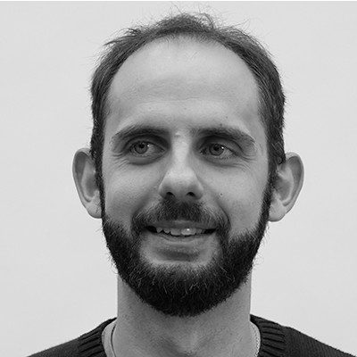 A black and white portrait of a man with short dark hair, beard and moustache wearing a dark t-shirt.