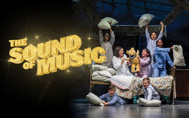 Maria and the Von Trapp children in pyjamas having a pillow fight on a bed, next to the shimmering gold title logo for The Sound of Music.