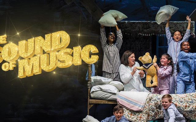 Maria and the von Trapp children in pyjamas having a pillow fight on a bed, next to the shimmering gold title logo for The Sound of Music.