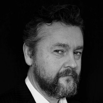 A black and white close-up of a man with short dark hair and goatee, wearing a white shirt and dark jacket.
