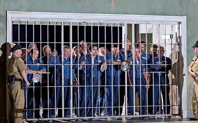 A group of prisoners behind a gate are secured by two guards as a man and woman on a balcony above overlook.