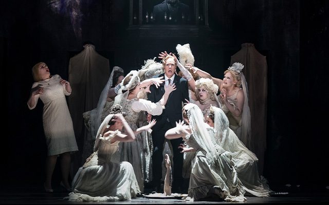 A man in a black suit stands surrounded by ghostly women wearing wedding dresses.