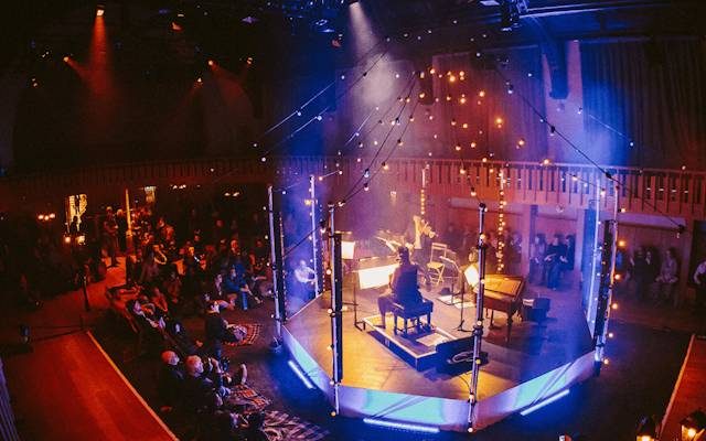 Musicians perform on a stage lit with fairy-lights as an audience sits watching.