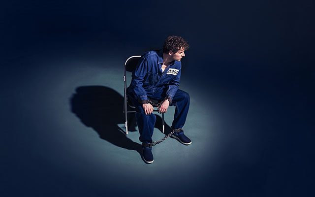 A man wearing a blue prison uniform sits in a chair in dark, atmospheric lighting.