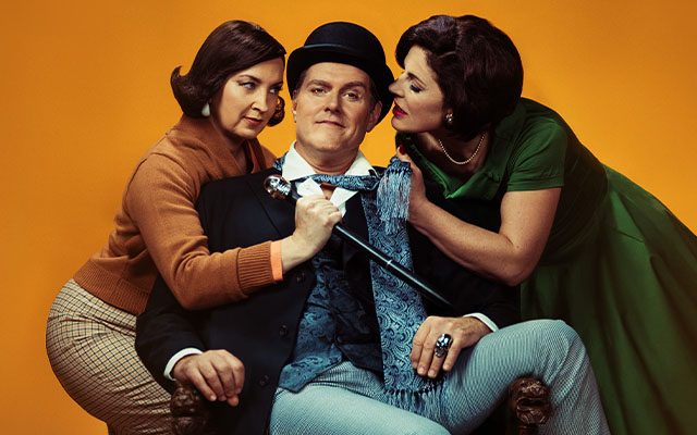 A man wearing a bowler hat sits with two women leaning into him.