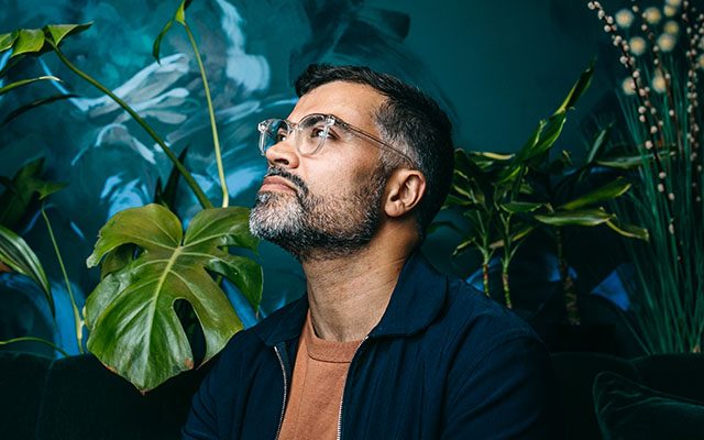 A man wearing glasses and a dark shirt stands in thought.