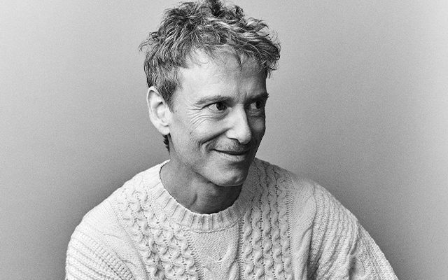 A man with short light hair, wearing a white jumper smiling.
