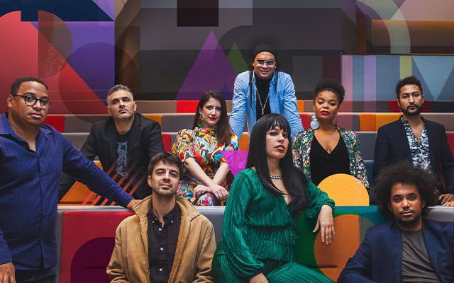 A group of ten people in stylish, artistic attire posing on tiered seating. The walls and furniture are decorated with vibrant, overlapping geometric patterns in a variety of colours.