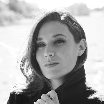 A black and white portrait of a woman with long dark hair, holding the collar of her coat outdoors.