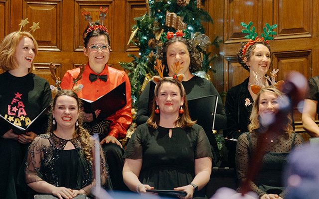 A group of chorus singers wearing antler hats and festive Christmas attire in song.