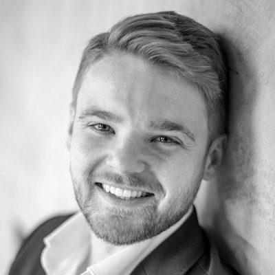 A black and white close-up of a man with short blonde hair and beard smiling.