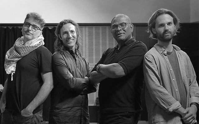A black and white portrait of four men standing in a row in a music studio, looking towards the camera.