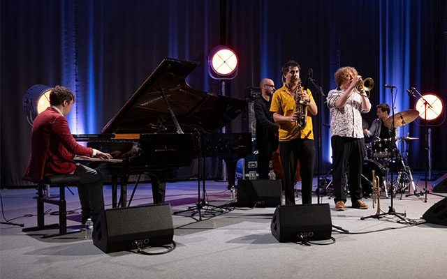 Five musicians playing instruments on an illuminated stage: piano, saxophone, trumpet, drums and double bass.
