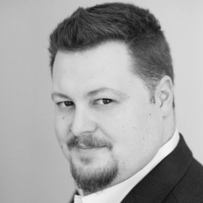 A black and white portrait of a man with short dark hair and a goatee beard, wearing a dark suit.