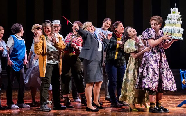 A conga line of of women dancing, carrying a large cake.