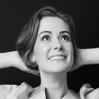 A black and white close-up of a woman with medium length dark hair resting her hands against the back of her head, smiling.