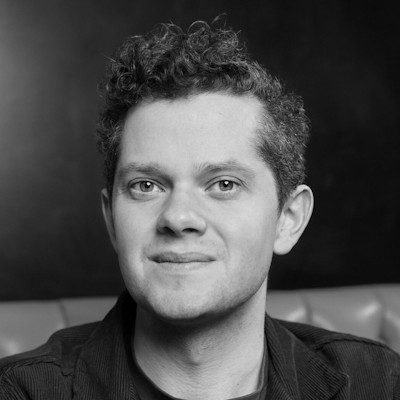 A black and white portrait of a clean-shaven man with short dark hair, wearing a dark t-shirt.