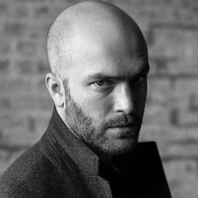 A black and white portrait of a man with a dark beard and clean-shaven head.