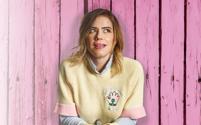 A woman with light-coloured long hair wearing a yellow jumper, sitting in front of pink wood panels.