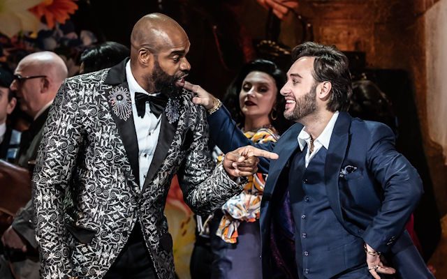 Two men in ostentatious suits speaking to each in an Opera North performance of Rigoletto.