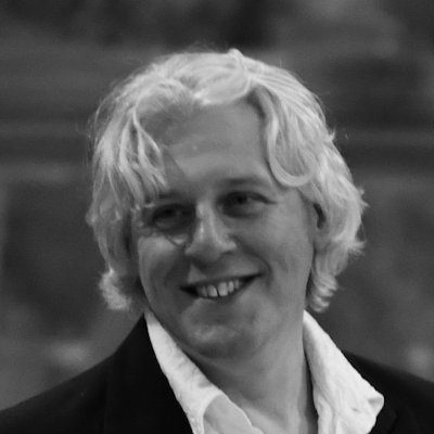 A black and white portrait of a man with long blonde hair smiling.