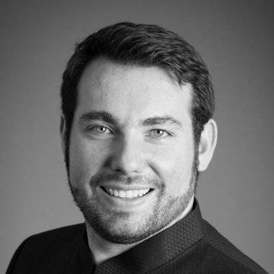A black and white portrait of a man with short dark hair and a short beard, wearing a dark suit, smiling at the camera.