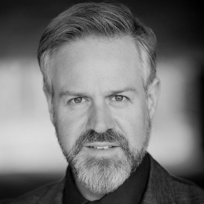 A black and white portrait of a man with short hair and a greying beard.
