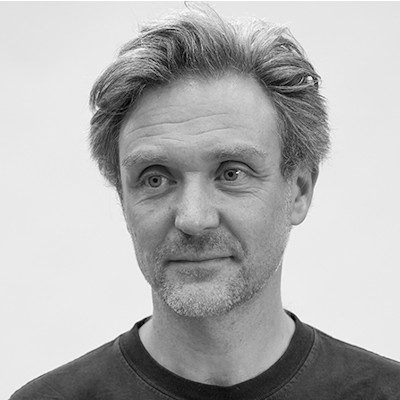 A black and white head-shot of a man with short greying hair, wearing a black t-shirt.