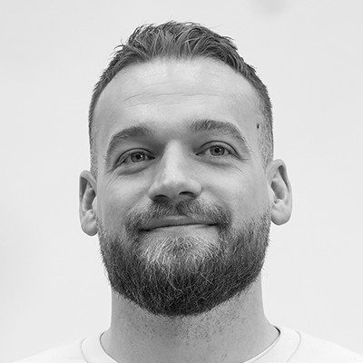A black and white close-up of a man with short dark hair, moustache and beard, wearing a white t-shirt.
