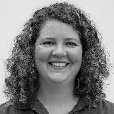 A black and white close-up of a woman with long curly hair wearing a dark t-shirt.