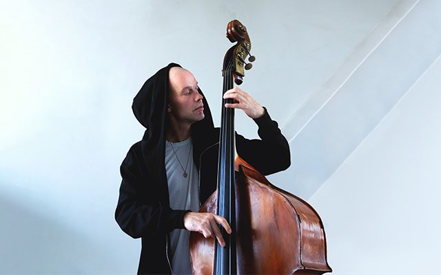 A man wearing a black hoodie and grey t-shirt playing a double bass.