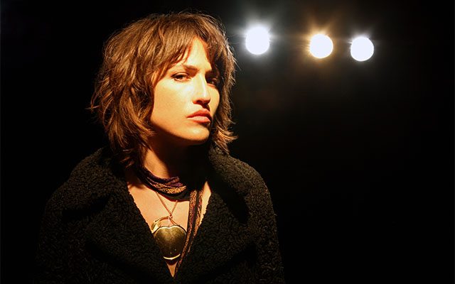 Joan As Police Woman looking directly at the camera, wearing a black textured coat and gold necklace, set against a dark background with three warm stage lights.