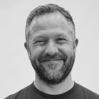 A black and white close-up of a man with short medium-coloured hair, moustache and beard, wearing a dark t-shirt.