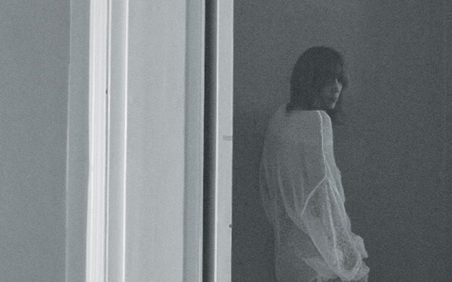 A grainy, black and white image of a woman with long dark hair, wearing a long white shirt.