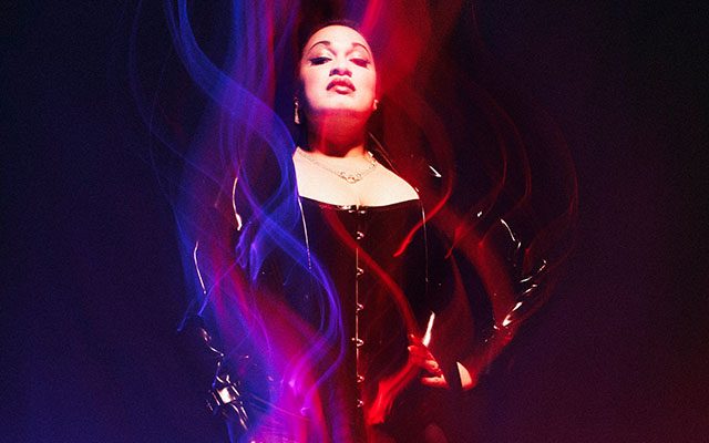 A woman posing in a dark setting, framed by vibrant, flowing streaks of red and blue light. They are wearing a dark corset and jacket, looking upward with a confident expression.