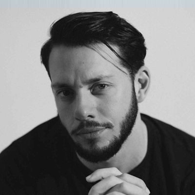 A black and white head-shot of Thomas Chenhall - a man with short dark hair and beard, wearing a black jumper.