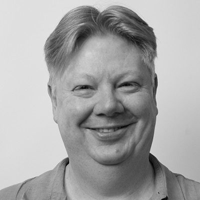 A black and white close-up of a man with short light hair, wearing a light t-shirt smiling.