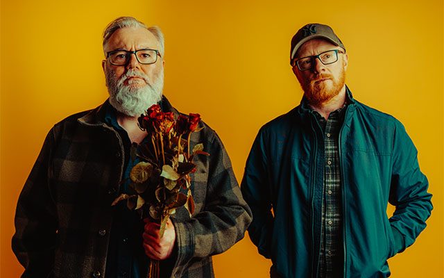 Two men standing, one with grey hair, glasses and a beard holding a bunch of flowers, the other with ginger hair, beard and baseball cap, in front of an orange wall.