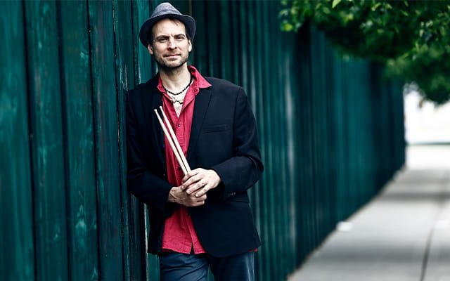 A man wearing a black suit jacket, red shirt and jeans leaning against a garden fence holding a pair of drum sticks.