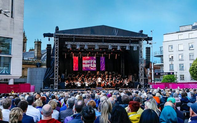 The orchestra of Opera North performing live on stage in Millennium Square, Leeds in front of a large crowd.