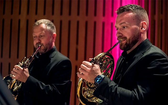 Two men playing horns on an stage in front of a wood background illuminated with colourful red light.